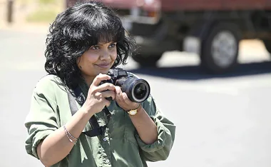 Miranda Tapsell, wearing a khaki top, is holding a camera and looking at someone.
