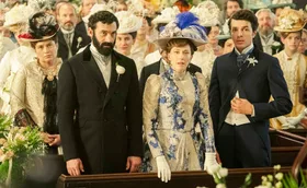 Two men in suits beside a woman dressed in a beautiful gown with an old fashioned hat.