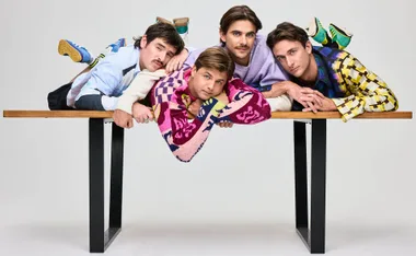 From left to right: Dom, Liam, Matt and Jack all lie on a table posing as they stare into the camera while wearing colourful shirts and jumpers