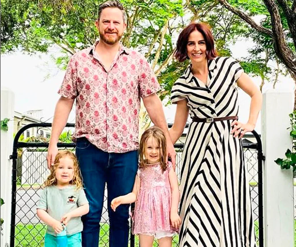 Alana is wearing a black and white stripe dress in front of a gate with greenery behind her as she poses with her husband and two kids.