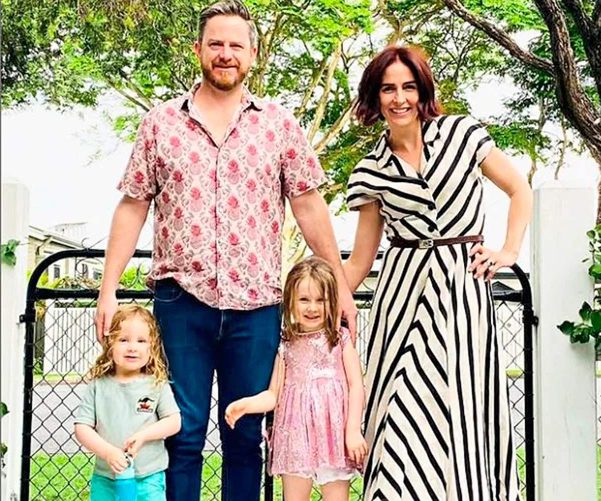 Alana is wearing a black and white stripe dress in front of a gate with greenery behind her as she poses with her husband and two kids.