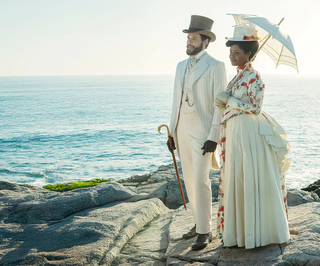 A man and woman are dressed in all white old fashioned attire, the woman with a parasol. They stand on a rock with the ocean in the background.