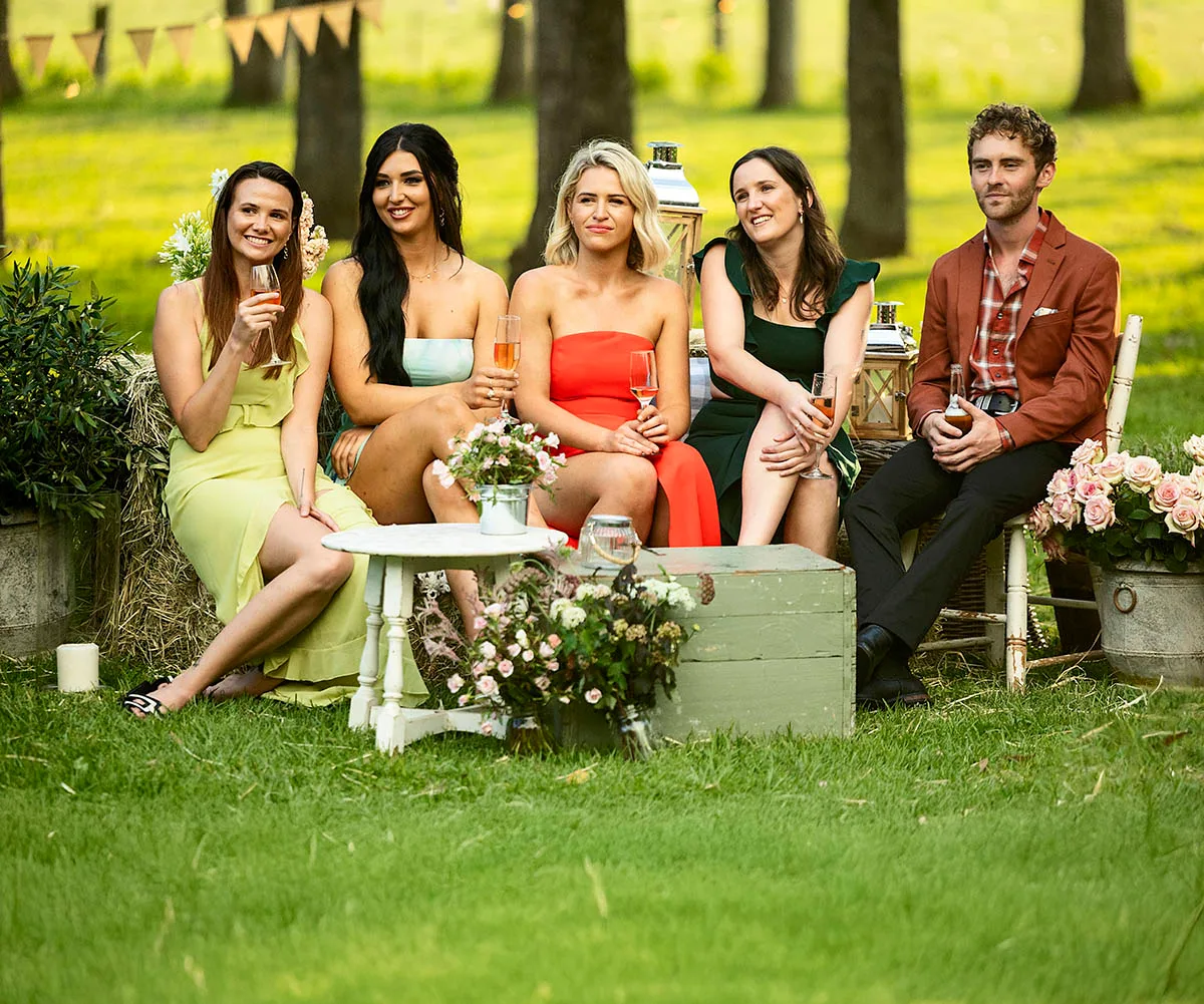 Farmer Jack (right) is pictured with four ladies of his farm all in beautiful gowns as they hold champagne glasses amongst a rustic setting of flowers, green grass, trees and little tables and chairs. 