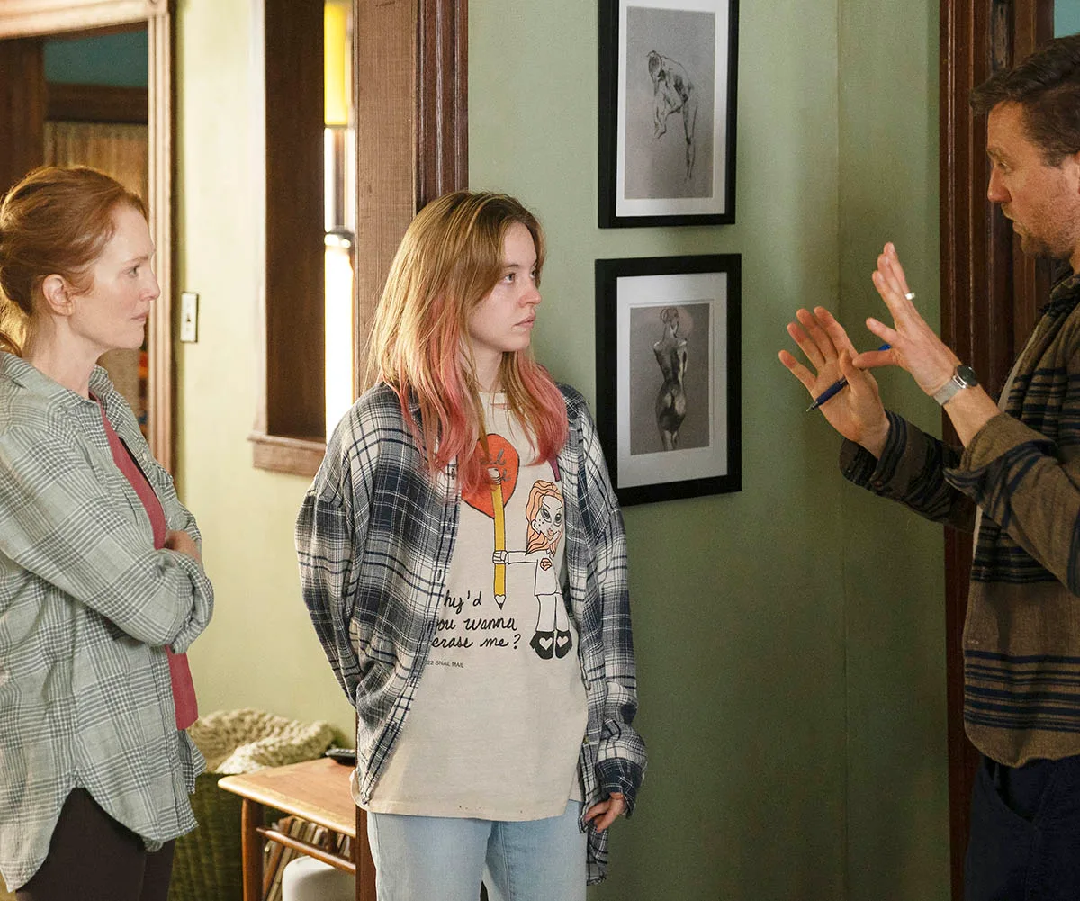 Julianne Moore and Sydney Sweeney stand on set, inside a house, while director Michael Pearce talks to them.