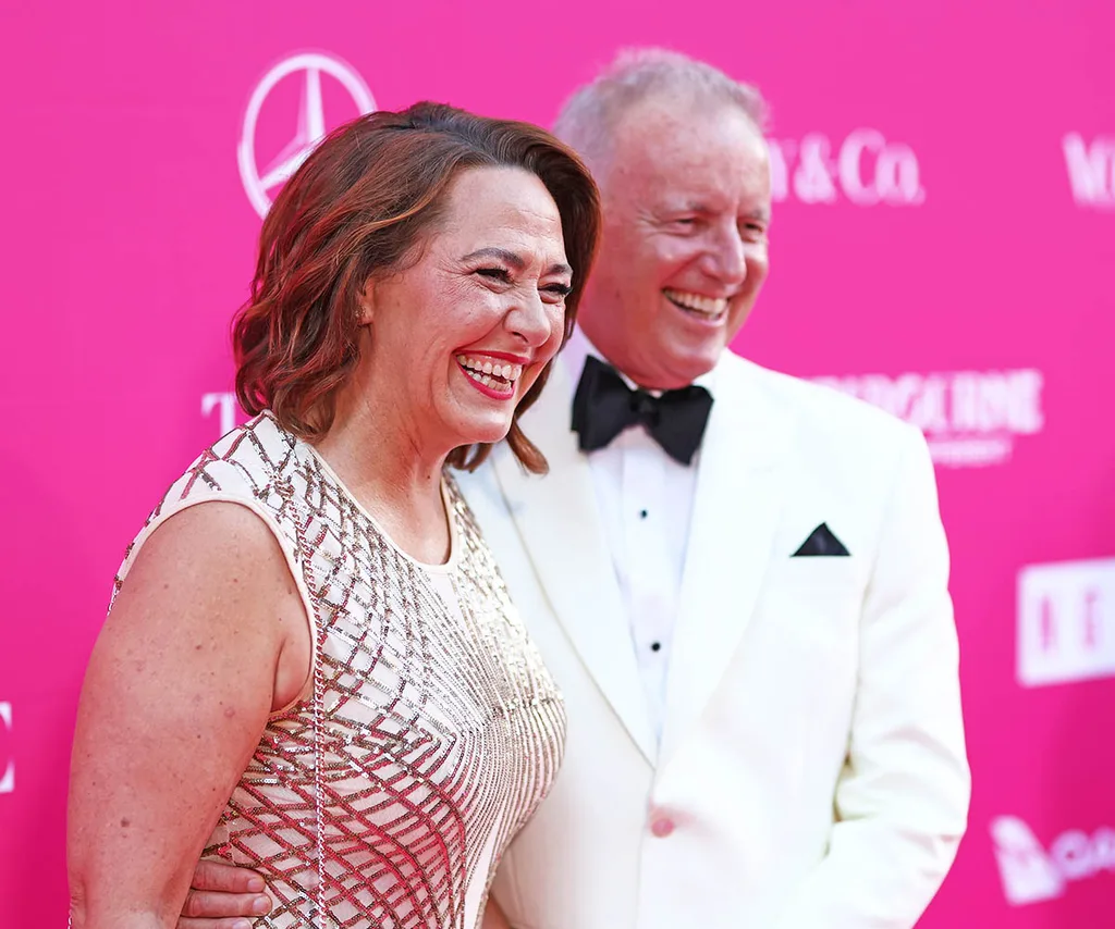 Lisa Millar, in a silver dress, and her partner Simon Carless, in a white suit and black bow tie, smile on the red carpet, in front of a pink background.