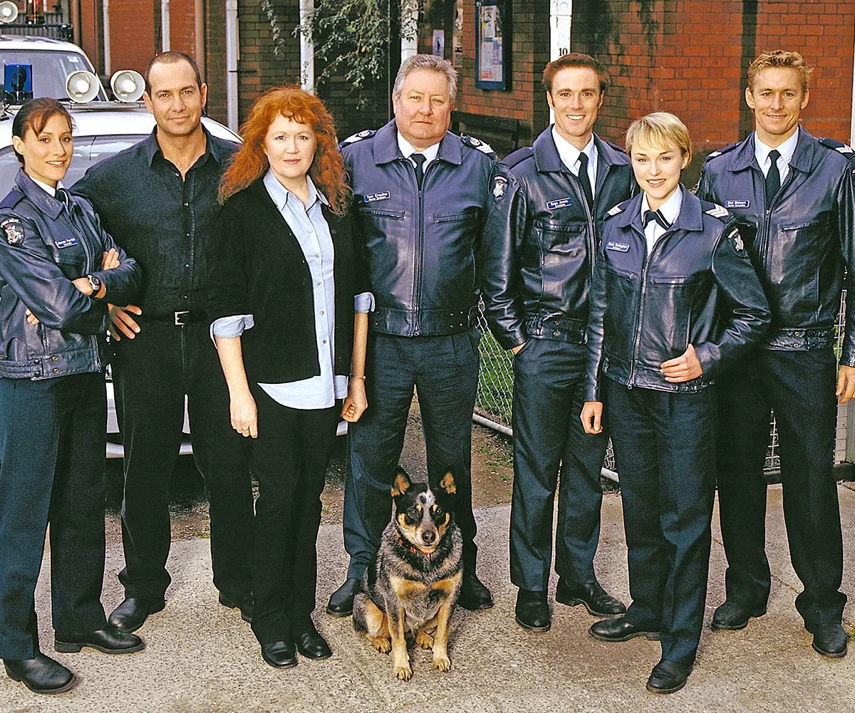 Martin Sacks (left) pictured with Blue Heelers cast all wearing their police uniform except for himself and the red haired woman to his right. A dog is pictured at their feet. 
