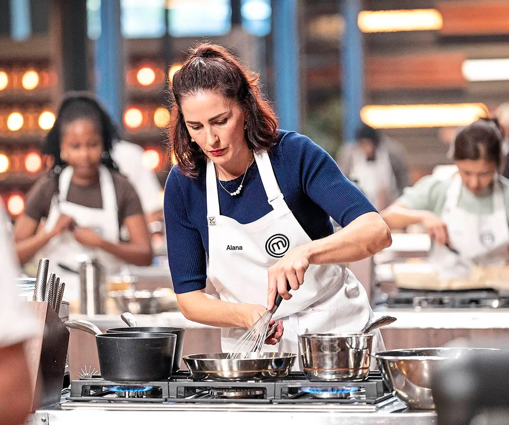 Alana focuses as she cooks in the MasterChef kitchen while wearing a navy top and a white MasterChef apron