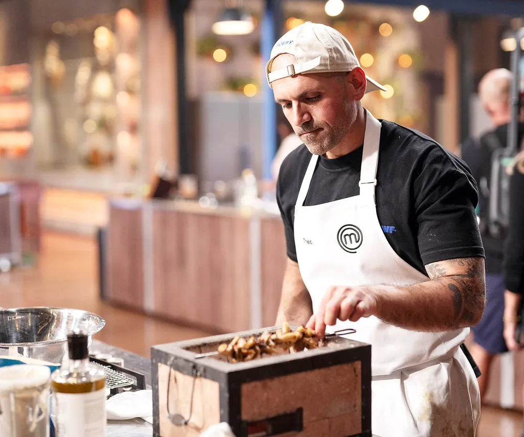Theo wears a black t-shirt, white backwards cap and a white apron as he cooks a skewer on a hibachi grill in the MasterChef kitchen