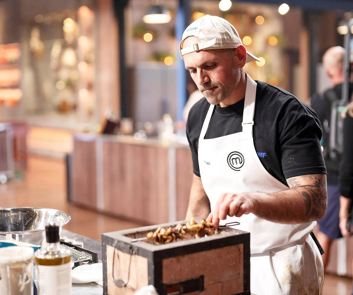 Theo wears a black t-shirt, white backwards cap and a white apron as he cooks a skewer on a hibachi grill in the MasterChef kitchen