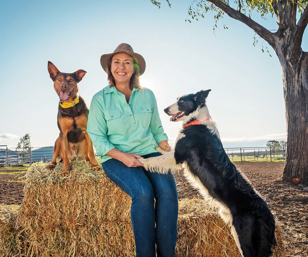 Lisa Millar sits on a bale of hay, in the countryside, with one dog's paws in her lap and another dog sitting next to her.