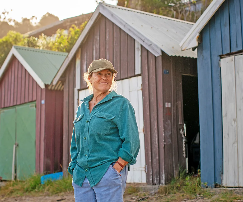 The Survivors. Robyn Malcolm as Verity in The Survivors. Verity is dressed casually in jeans and a loose work style shirt with a cap on in front a barn.