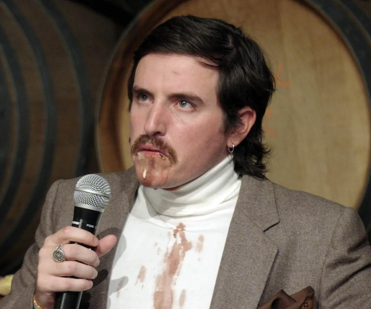 Dom is pictured in a white turtle neck, beige suit jacket holding a microphone with food and wine all over his chin and down his shirt