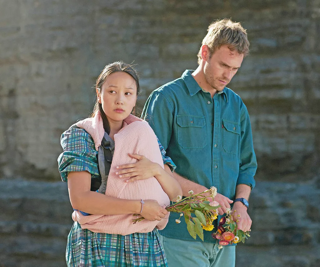 Yerin Ha as Mia, Charlie Vickers as Kieran in The Survivors. Mia is holding their baby in a pink blanket and flowers while walking beside Kieran while they both look sad.