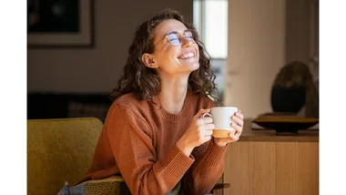 A smiling woman drinks a hot beverage with a blanket on her lap.