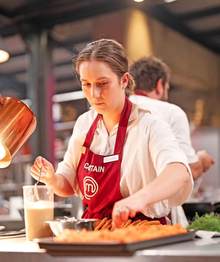 Laura wears a white button up shirt and a red MasterChef apron as she cooks in the kitchen looking very focused.