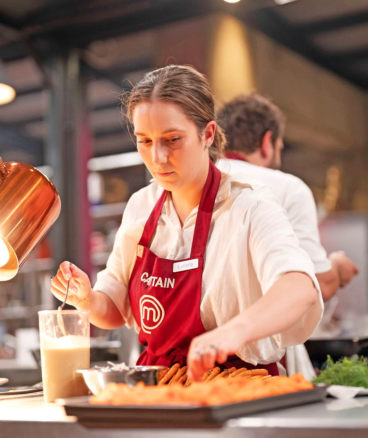 Laura wears a white button up shirt and a red MasterChef apron as she cooks in the kitchen looking very focused.