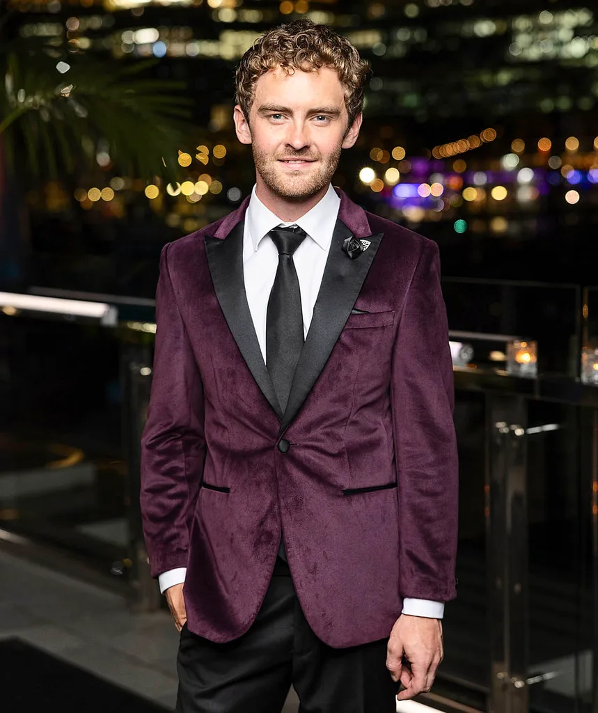 Farmer Jack is pictured on a balcony at night-time with the city skyline in the back drop. He wears a purple velvet suit jacket, white shirt and black tie and suit pants.