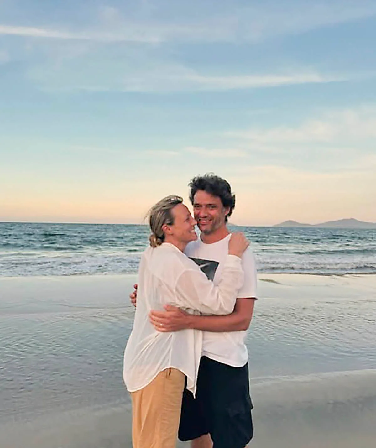 Marta and her husband Ben, both wearing white shirts, hold each other as the sun sets on a beach behind them.