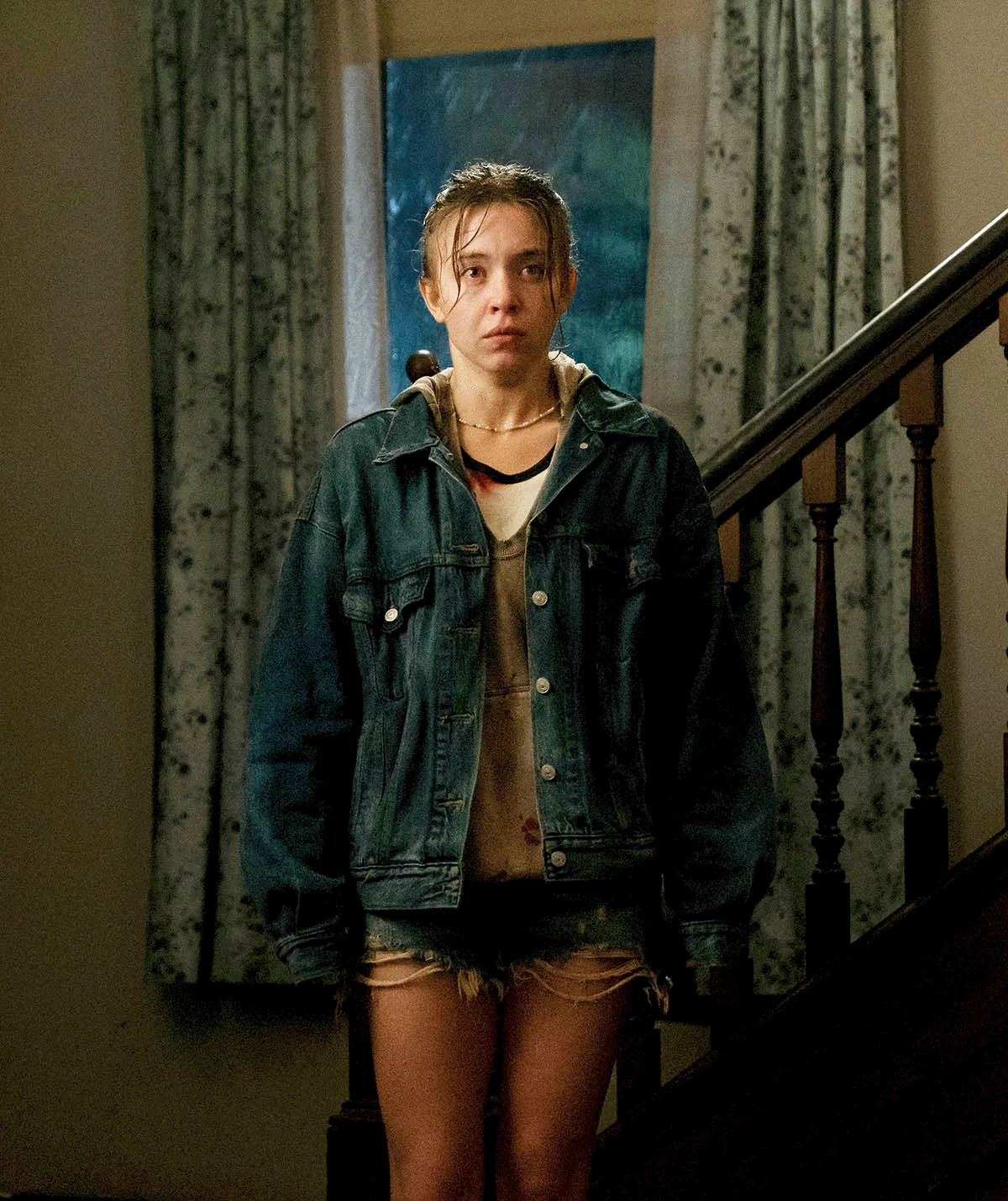 Sydney Sweeney, looking scruffy, stands near the bottom of a staircase in a scene from Echo Valley. 
