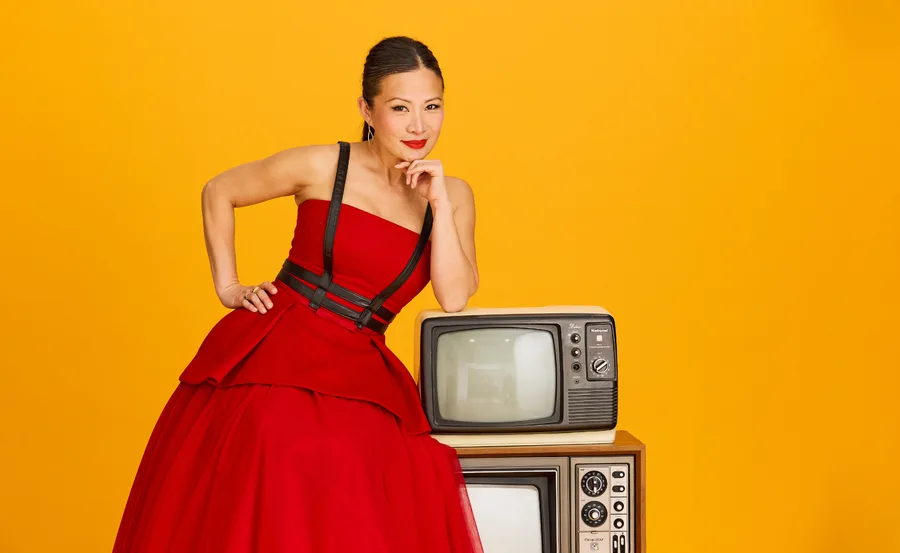 Poh, dressed in a red and black dress, leans on two old TV sets, against an orange background
