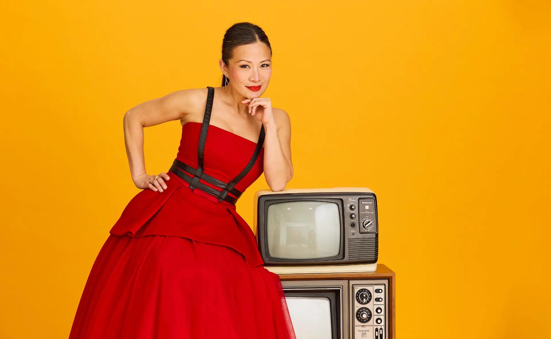 Poh, dressed in a red and black dress, leans on two old TV sets, against an orange background