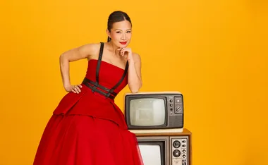 Poh, dressed in a red and black dress, leans on two old TV sets, against an orange background