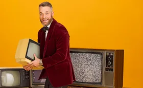 Hamish Blake in a maroon blaker, bowtie holding an old TV in front of three old TV's and a mustard backdrop - grinning.