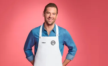 Callum wears a blue button up, white MasterChef apron and smiles as he poses in front of a pink backdrop.