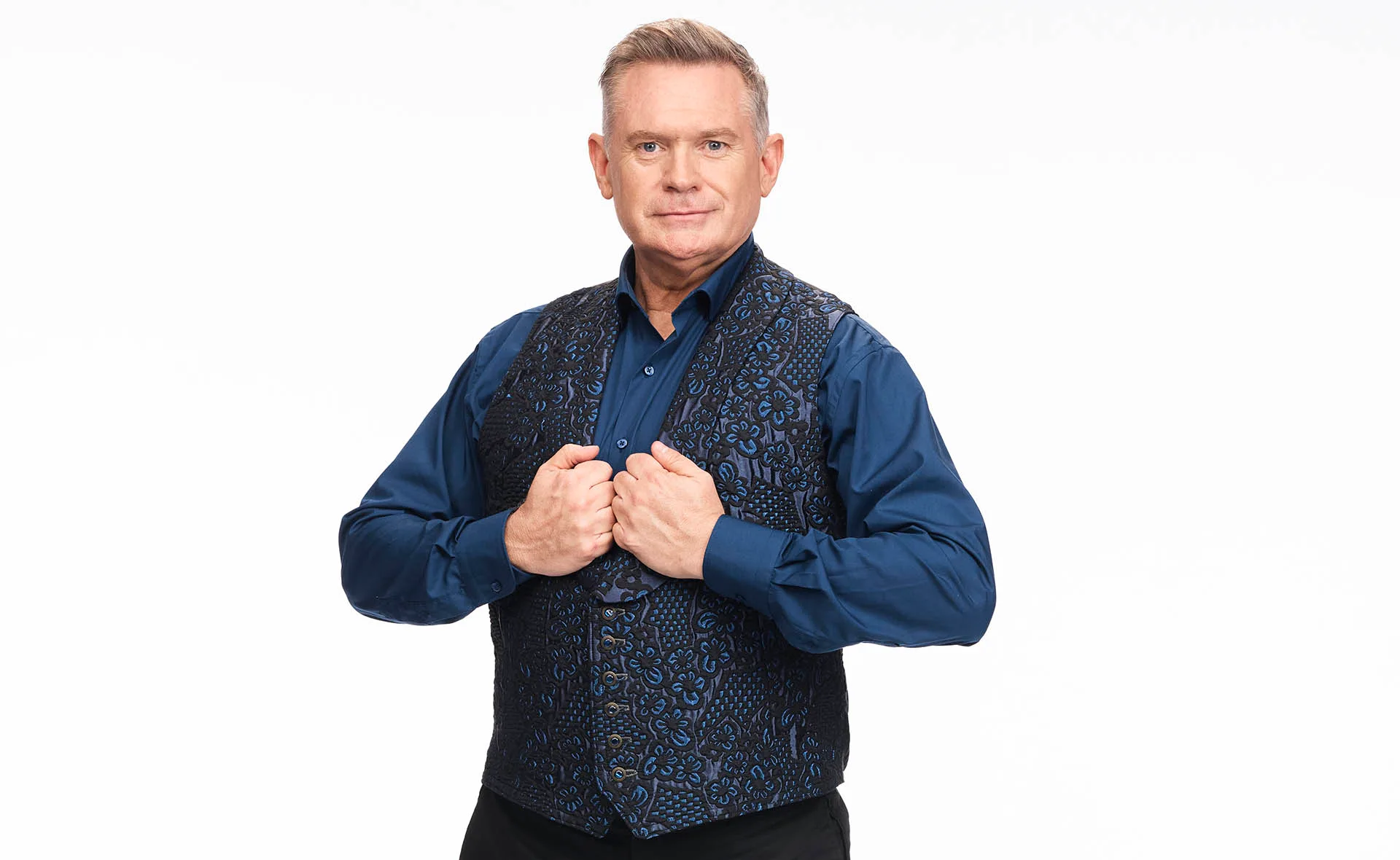 Michael Usher in a blue button up shirt with a matching navy vest. He clutches at the buttons as he poses against a white backdrop.