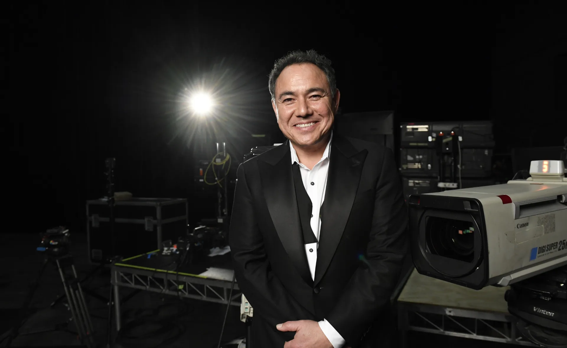 Sam Pang in a tuxedo with the bow undone. He stands in front of a back backdrop, a spotlight over his left shoulder and camera equipment behind him.