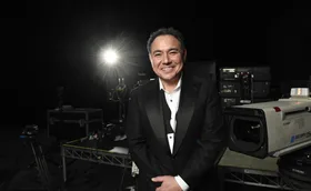 Sam Pang in a tuxedo with the bow undone. He stands in front of a back backdrop, a spotlight over his left shoulder and camera equipment behind him.