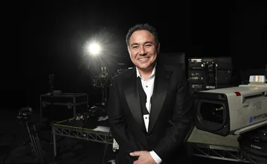 Sam Pang in a tuxedo with the bow undone. He stands in front of a back backdrop, a spotlight over his left shoulder and camera equipment behind him.