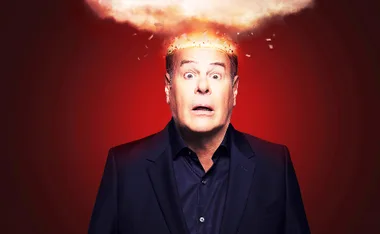 Dan is pictured in a black suit in front of a red background. He is open mouthed appearing shocked as his head looks like it is on fire.