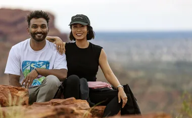 Melissa Leong sits next to Nazeem Hussain, her arm on his shoulder, on a rock, with a view of central Australia behind them.