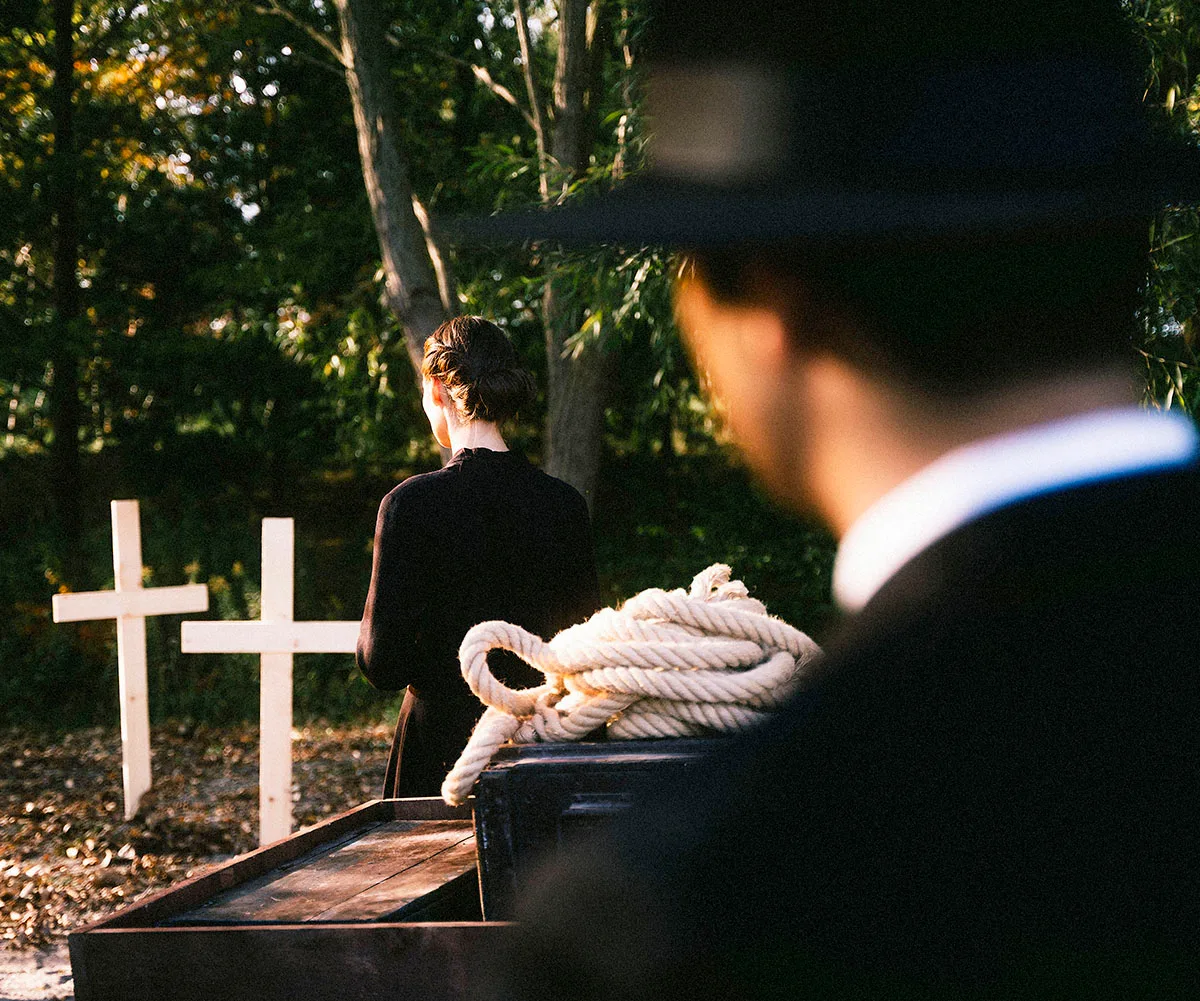 A woman looks to a white cross in the ground in a graveyard. A man with a hat and a suit on watches on, blurry in the foreground.