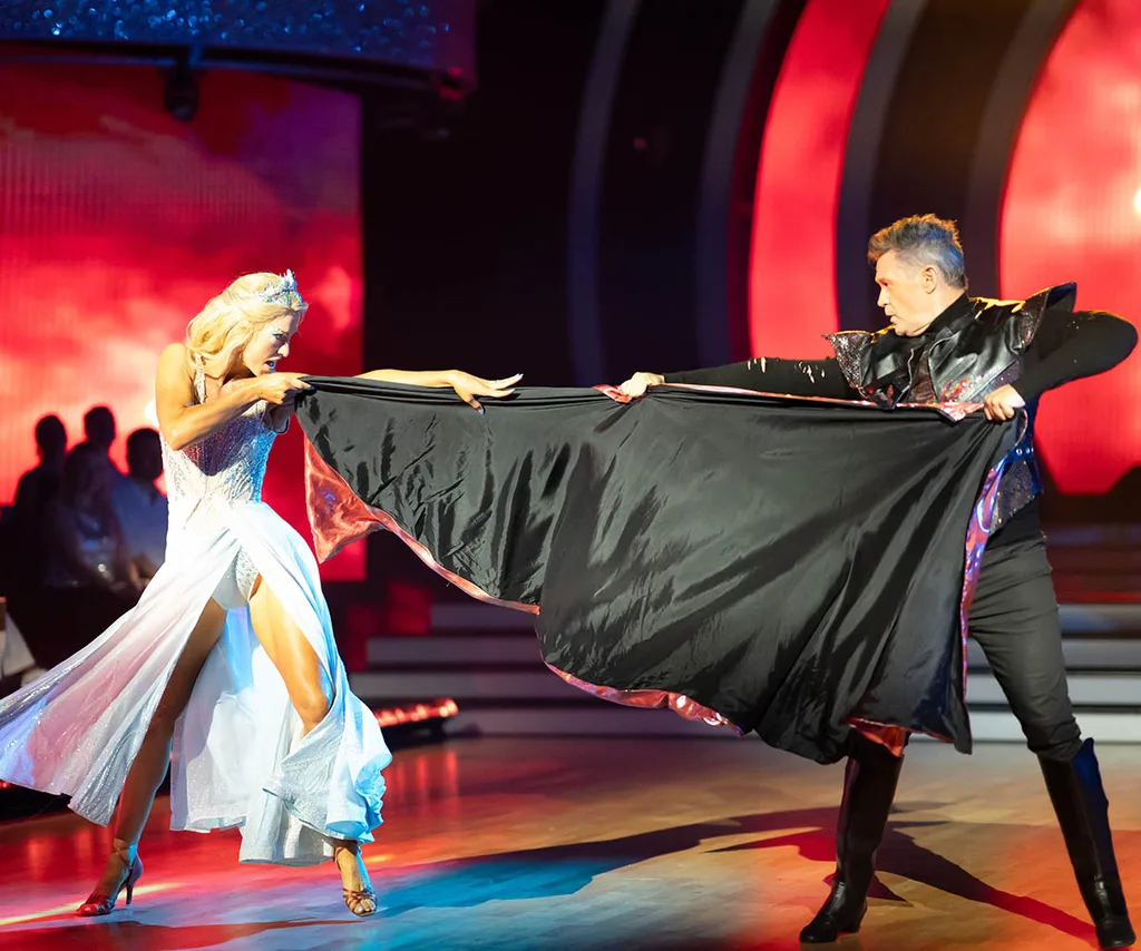 Natalie and Michael on stafe both holding a piece of costume and pulling it between them! She wears a blue dress with a tiara and he is in a black matador style costume.
