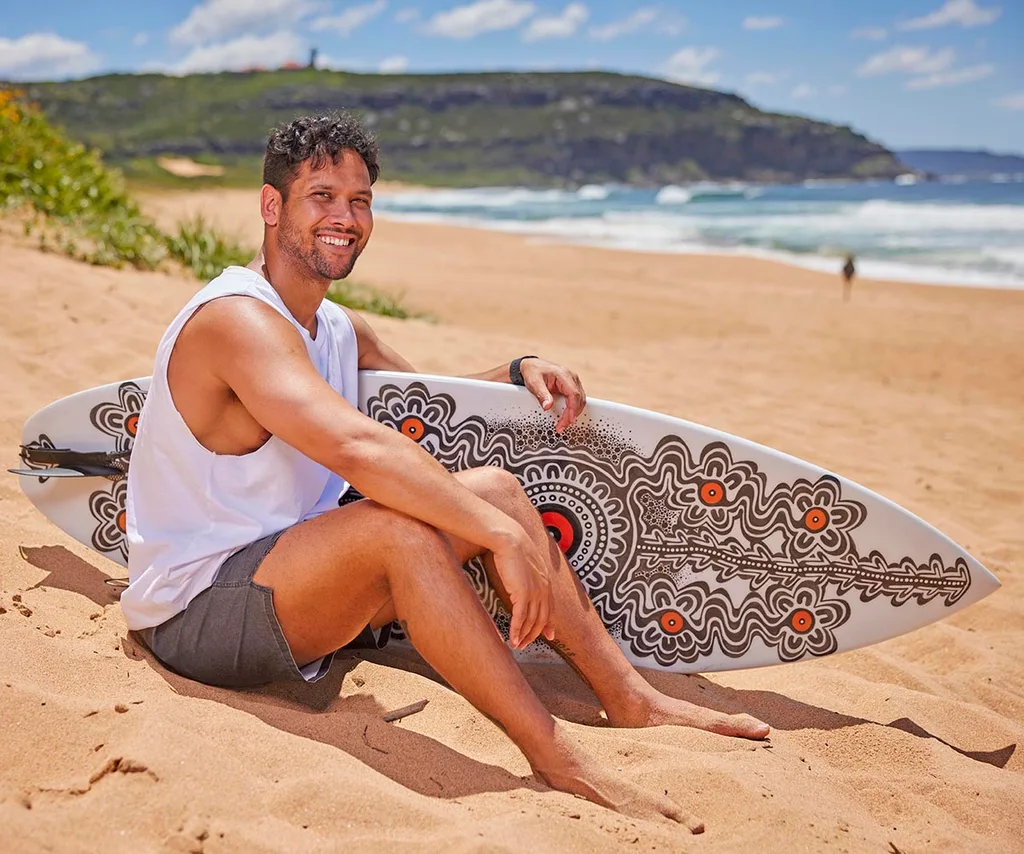 Kyle Shilling, as Mali Hudson, sits on the Summer Bay beach with a surfboard by his side.