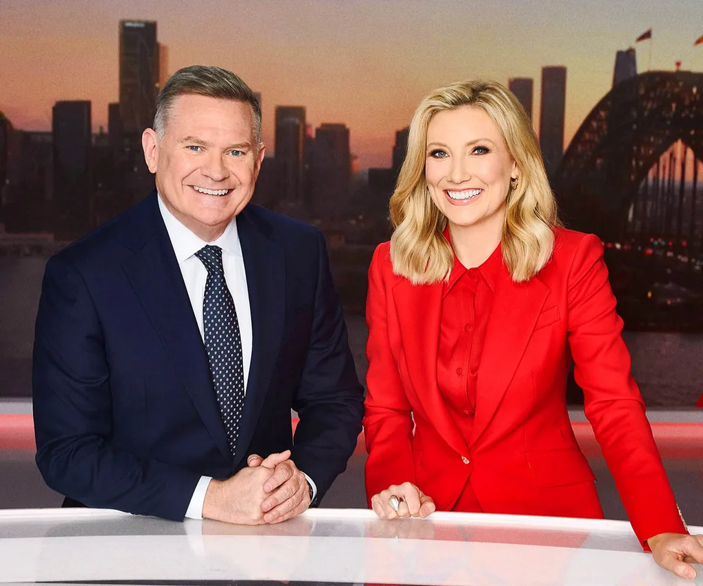 Michael in a suit and Angie in a red power suit smiling at the 7NEWS desk with an image of Sydney skyline in the backdrop.