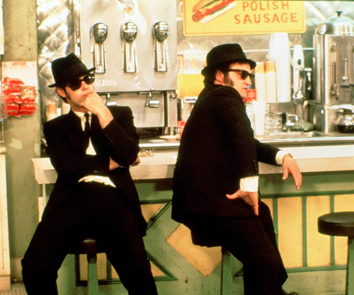 Dan Aykroyd and John Belushi in The Blues Brothers sitting at a counter in a diner with their iconic black and white suits, black sun glasses and hat. A sign in the background said 'Polish sausage'. 