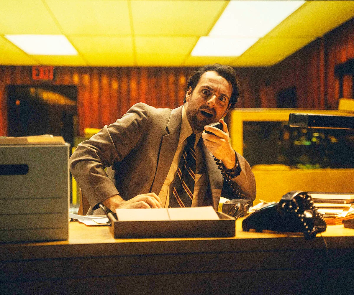 Man sits at a desk pouring over files in a brown box while on an old school phone taking a call looking shocked.