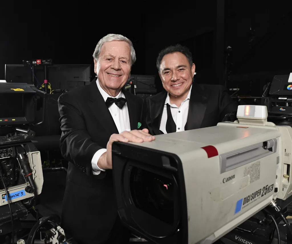 Sam Pang in a tuxedo with the bow undone to the left of Ray Martin in a tux. They stands in front of a back backdrop, a spotlight over his left shoulder and camera equipment behind him.