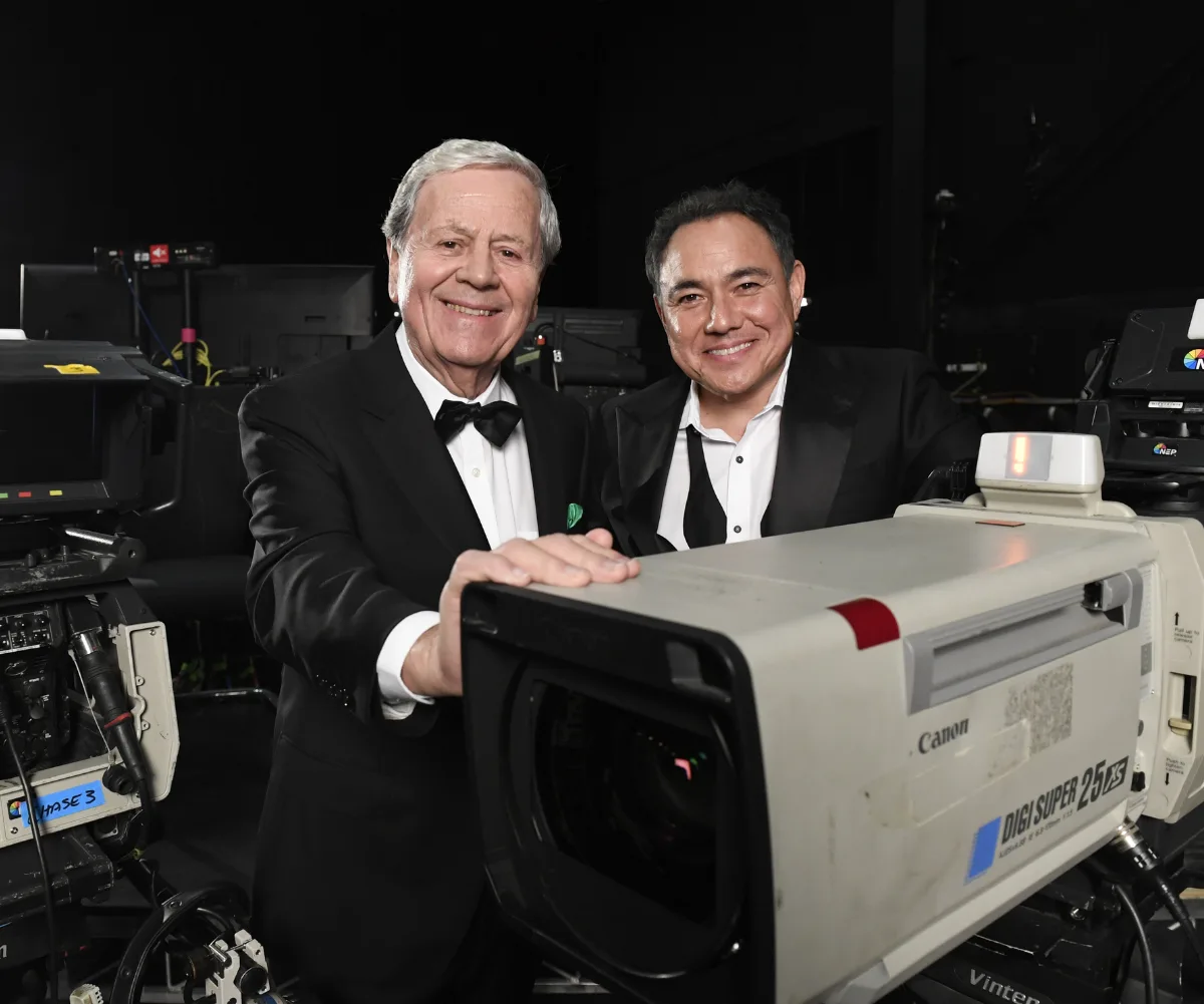 Sam Pang in a tuxedo with the bow undone to the left of Ray Martin in a tux. They stands in front of a  back backdrop, a spotlight over his left shoulder and camera equipment behind him.
