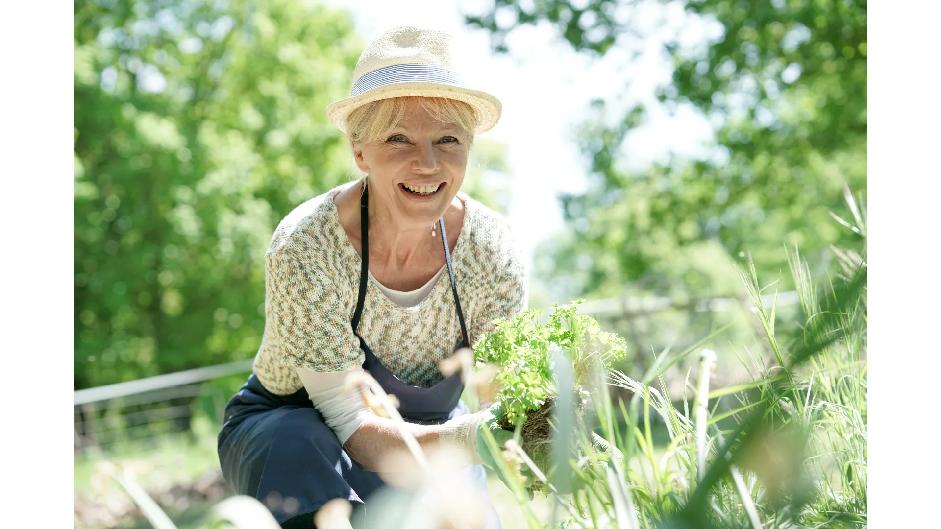 A blonde woman in a hat and apron smiles while gardening.