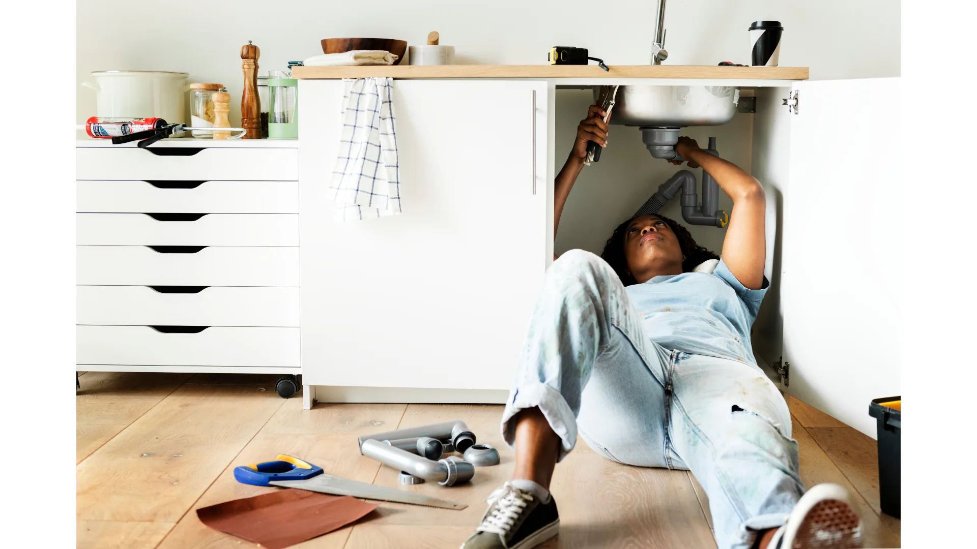 A woman repairs a pipe underneath a kitchen sink.
