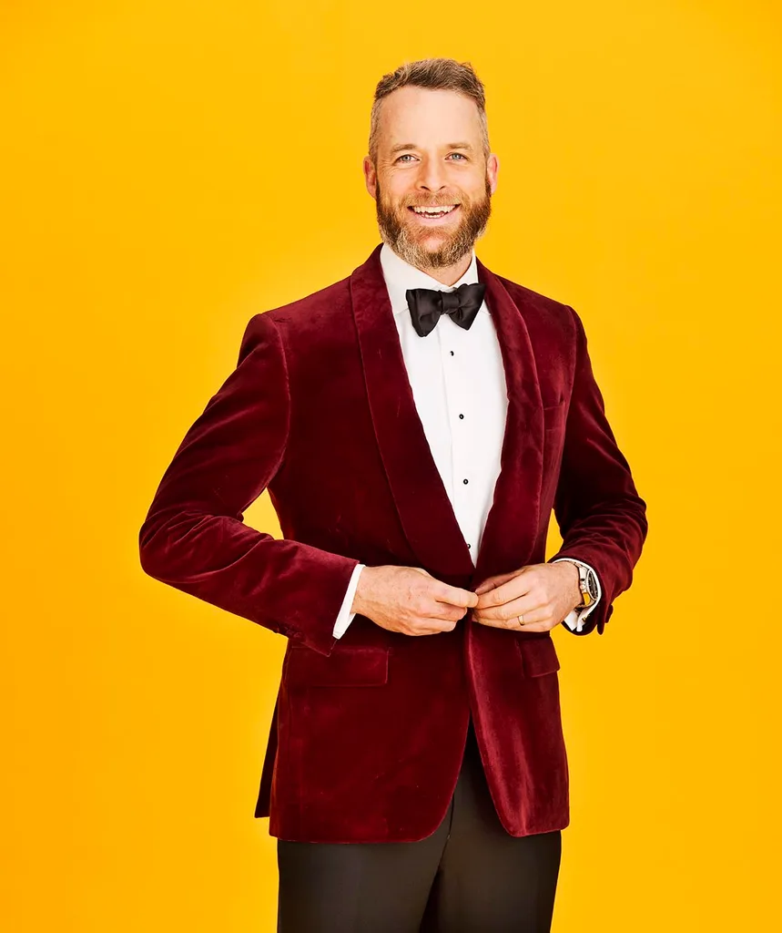 Hamish Blake smiling as he buttons up his maroon blazer, smiling against a mustard backdrop.