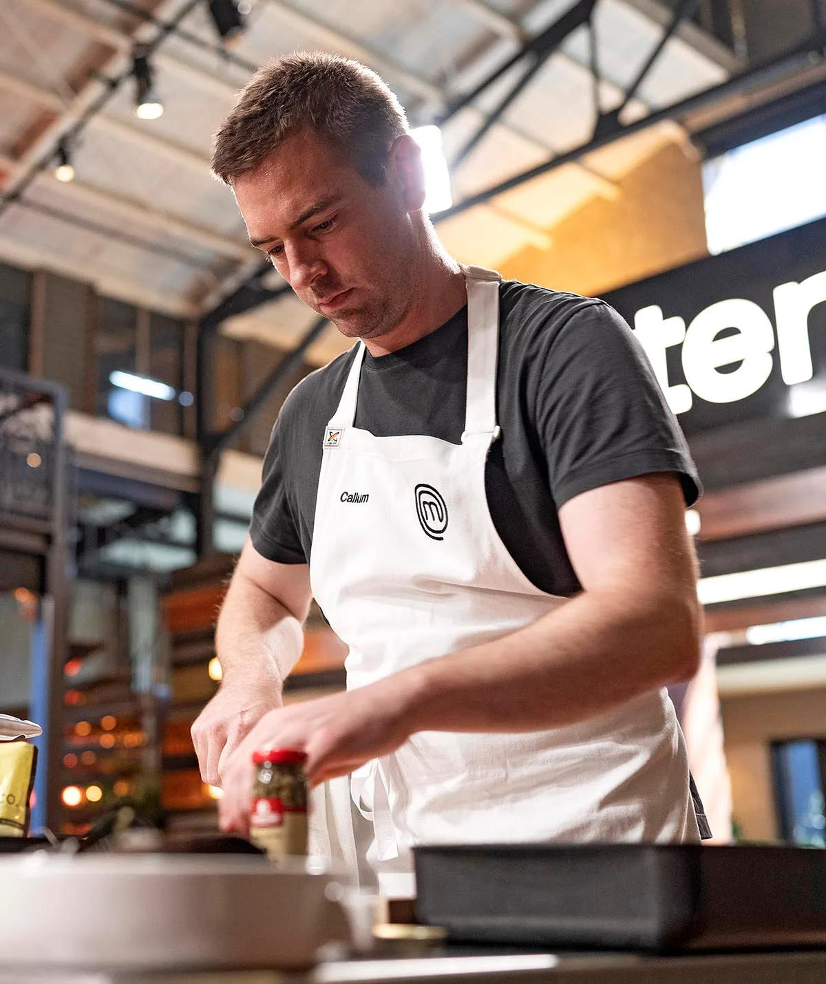Callum concentrates as he chops something in the MasterChef kitchen while wearing a black t-shirt and white MasterChef apron.