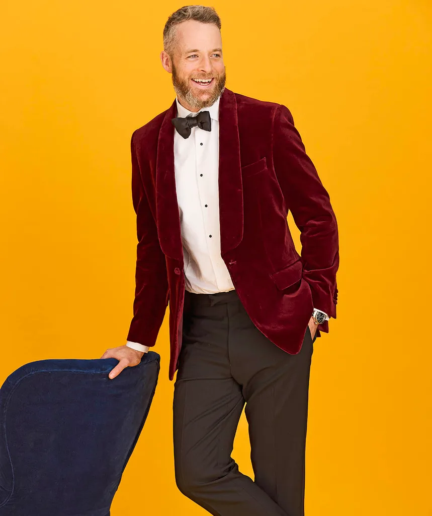 Hamish poses while holding a navy chair. He wears a maroon blazer, white shirt, black bow tie and stands against a mustard backdrop.