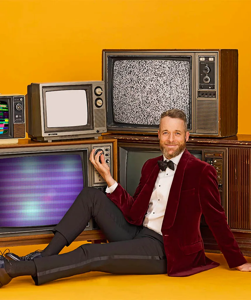 Hamish on the floor in front of a stack of old TV's, smiling as he spins the dial on one.