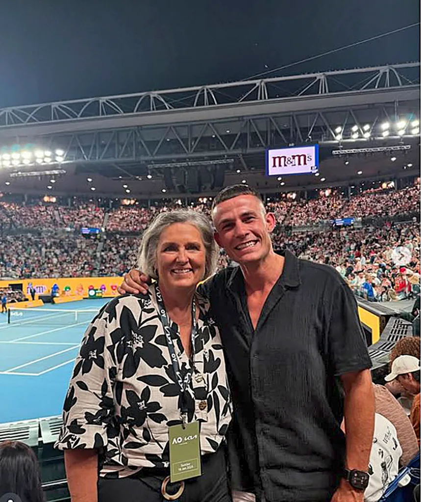 Kate on the left in a floral shirt and a lanyard around her neck and her son Harry smiling in a black shirt and his arm around her as they pose in front of a tennis match and full stadium.