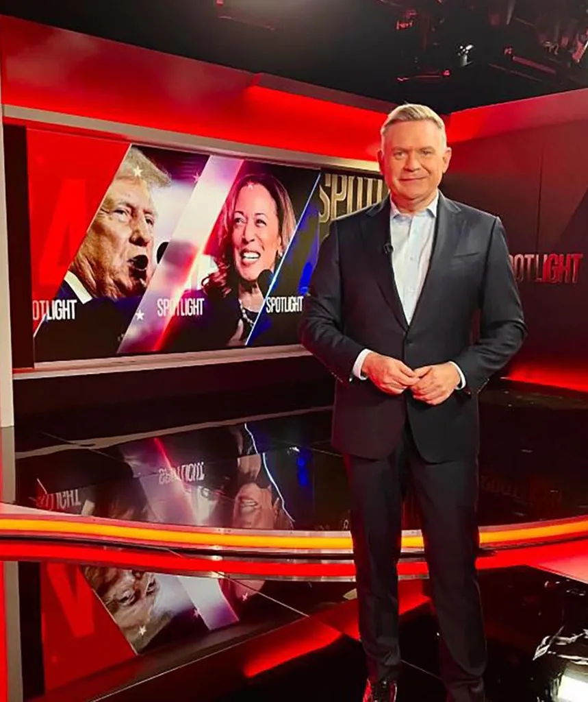 Michael poses in a black suit and white shirt on set for Spotlight with images of Donald Trump and Kamala Harris in the background.